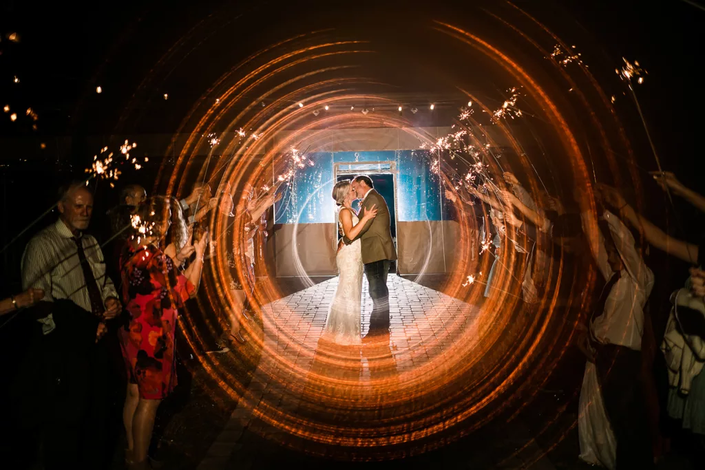 A couple in wedding attire kiss during a sparkler exit.